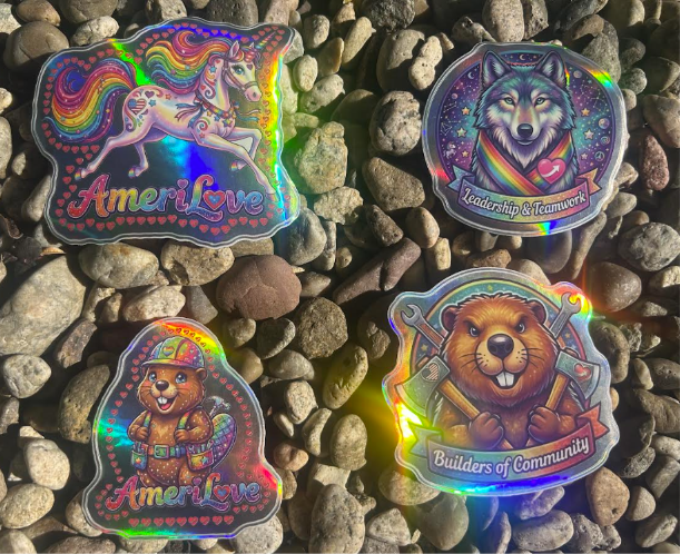 Four holographic America Learns sticker designs — AmeriLove unicorn, Leadership & Teamwork wolf, AmeriLove beaver, and Builders of Community beaver — arranged on river stones.